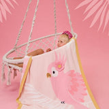 Child sitting in a hammock with a pink parrot-themed towel draped over them on a pink background.