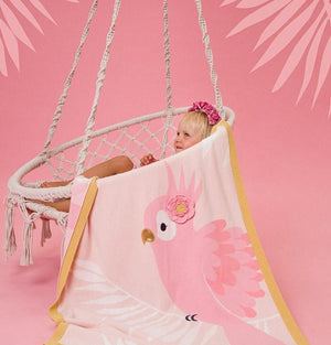Child sitting in a hammock with a pink parrot-themed towel draped over them on a pink background.