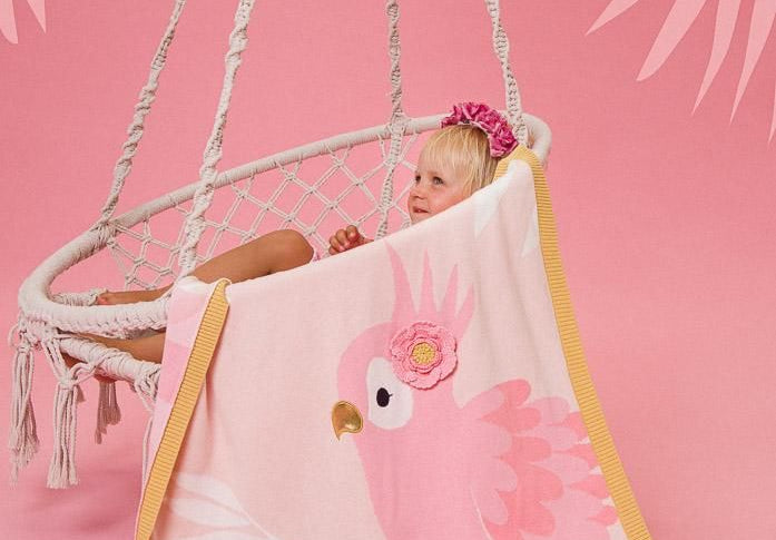 Child sitting in a hammock with a pink parrot-themed towel draped over them on a pink background.