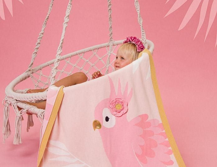 Child sitting in a hammock with a pink parrot-themed towel draped over them on a pink background.