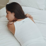 A woman lying on a white bed wearing a white ribbed tank top with a small black 'RE:DOPE' logo on the upper back.