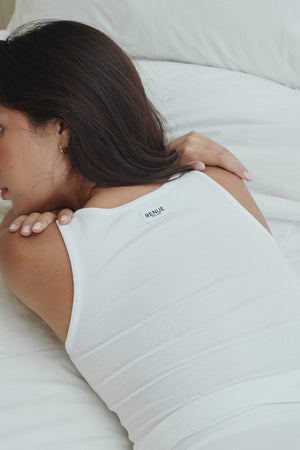 A woman lying on a white bed wearing a white ribbed tank top with a small black 'RE:DOPE' logo on the upper back.