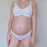 Pregnant model wearing white maternity bra and matching underwear against a plain light gray wall.