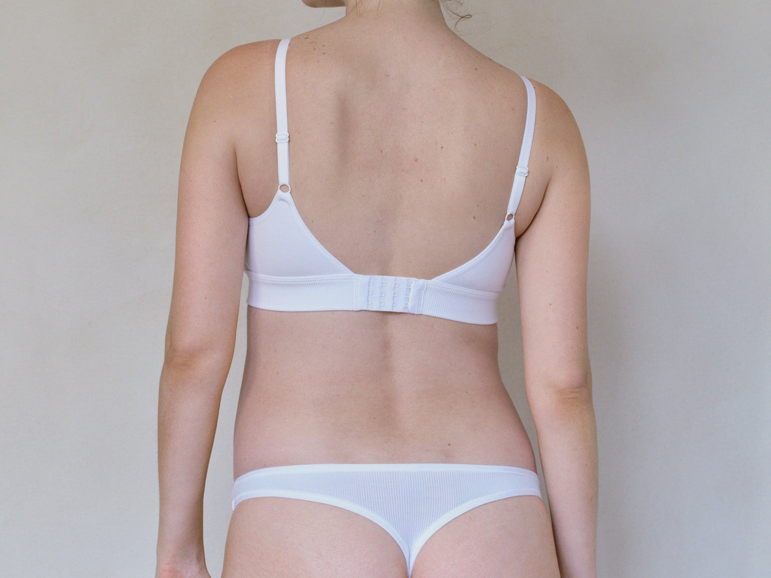 A woman shown from the back wearing a white bra and matching white underwear against a plain light gray wall.