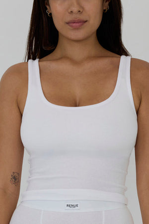 A woman wearing a white ribbed scoop-neck tank top and matching white bottoms, shown from the waist up against a light gray studio background. The brand name 'RENUE' is visible on the waistband label.