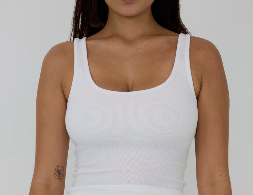 A woman wearing a white ribbed scoop-neck tank top and matching white bottoms, shown from the waist up against a light gray studio background. The brand name 'RENUE' is visible on the waistband label.