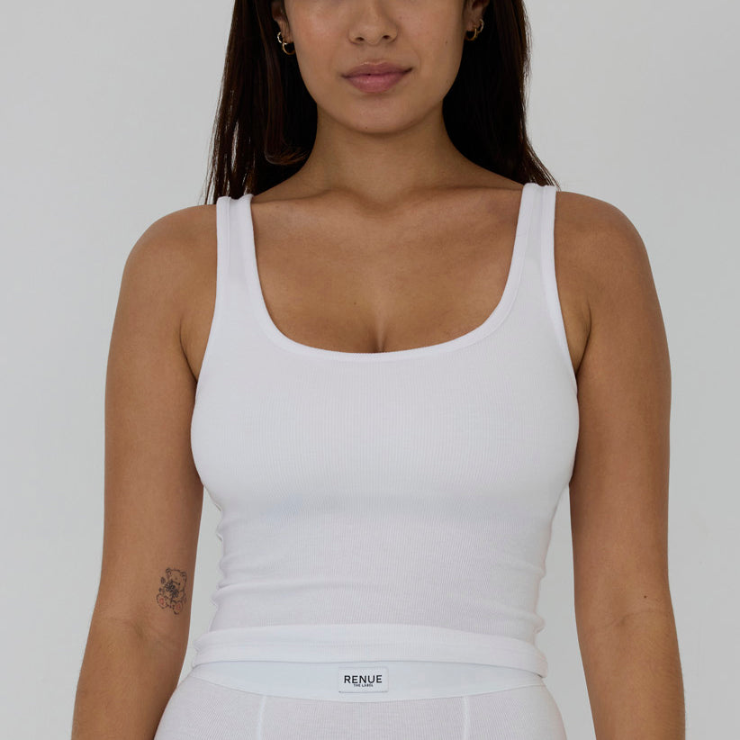 A woman wearing a white ribbed scoop-neck tank top and matching white bottoms, shown from the waist up against a light gray studio background. The brand name 'RENUE' is visible on the waistband label.