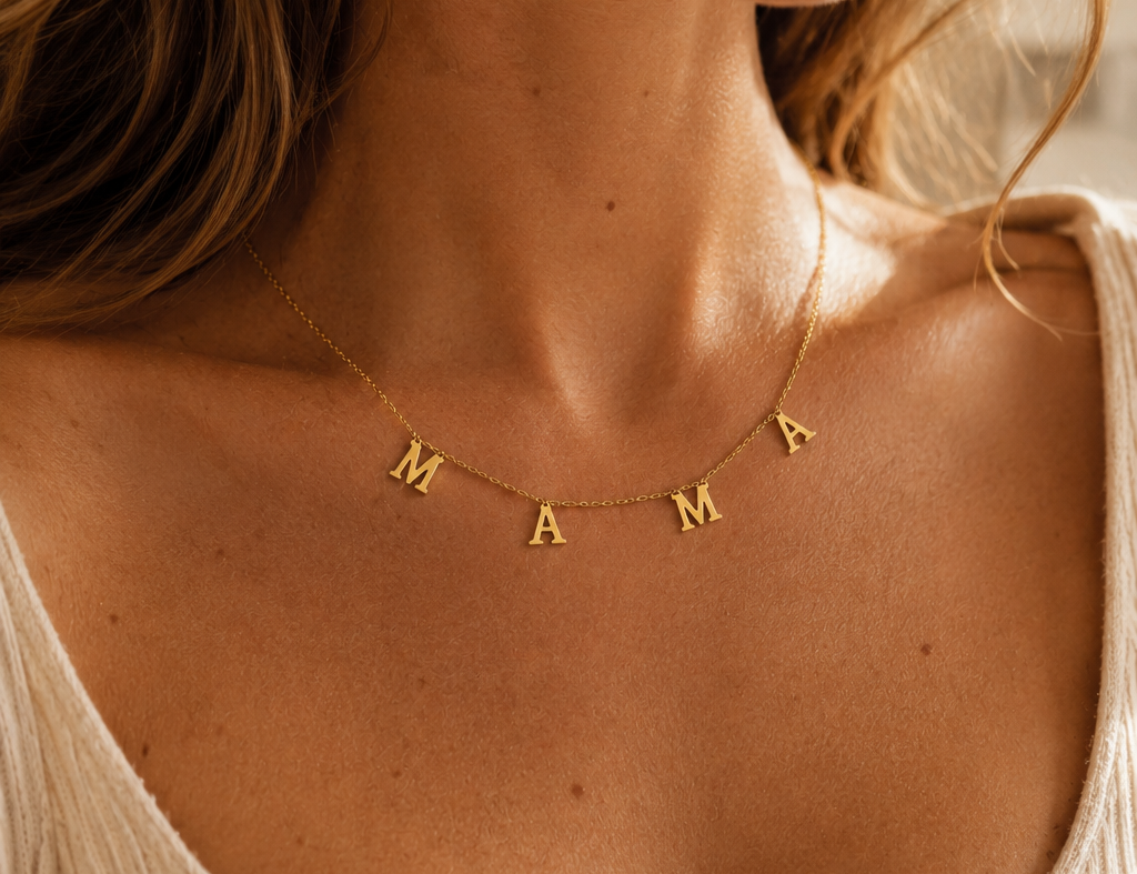 A close-up lifestyle shot of a woman wearing a delicate gold necklace with the name 'MAMA' spelled out in individual letter charms, styled with a beige ribbed scoop-neck top.