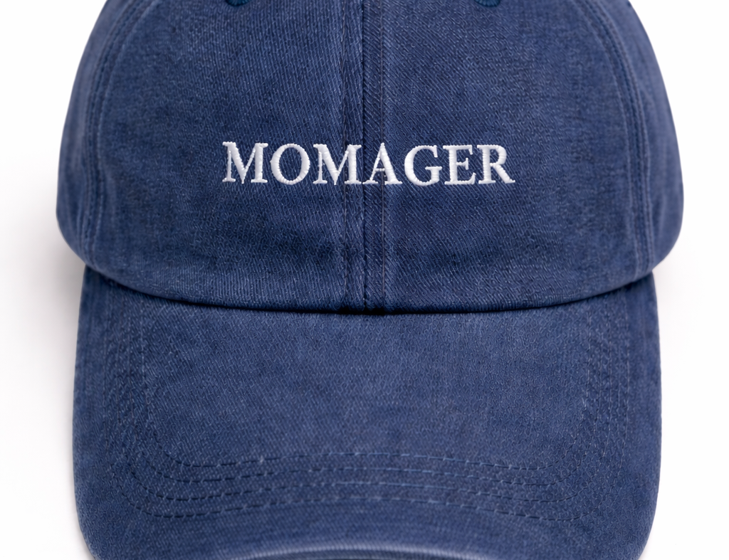 A navy blue washed cotton baseball cap with the word 'MOMAGER' embroidered in white serif letters across the front, shown against a plain white background.