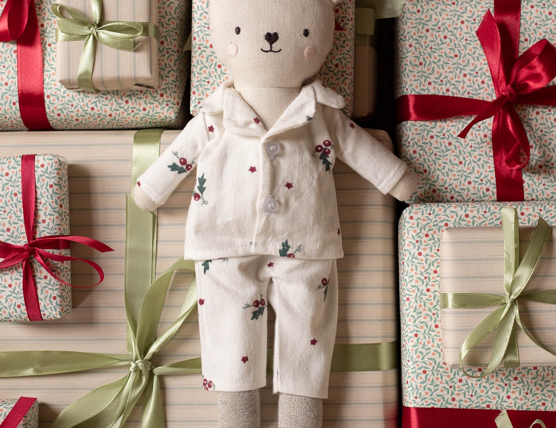 A plush teddy bear doll wearing cream-colored floral pajamas lies centered among wrapped gift boxes with red and green ribbons on a striped background.