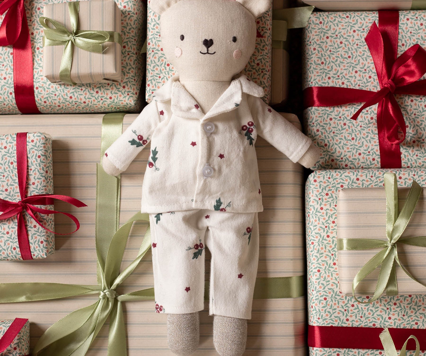 A plush teddy bear doll wearing cream-colored floral pajamas lies centered among wrapped gift boxes with red and green ribbons on a striped background.