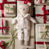 A plush teddy bear doll wearing cream-colored floral pajamas lies centered among wrapped gift boxes with red and green ribbons on a striped background.