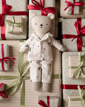 A plush teddy bear doll wearing cream-colored floral pajamas lies centered among wrapped gift boxes with red and green ribbons on a striped background.