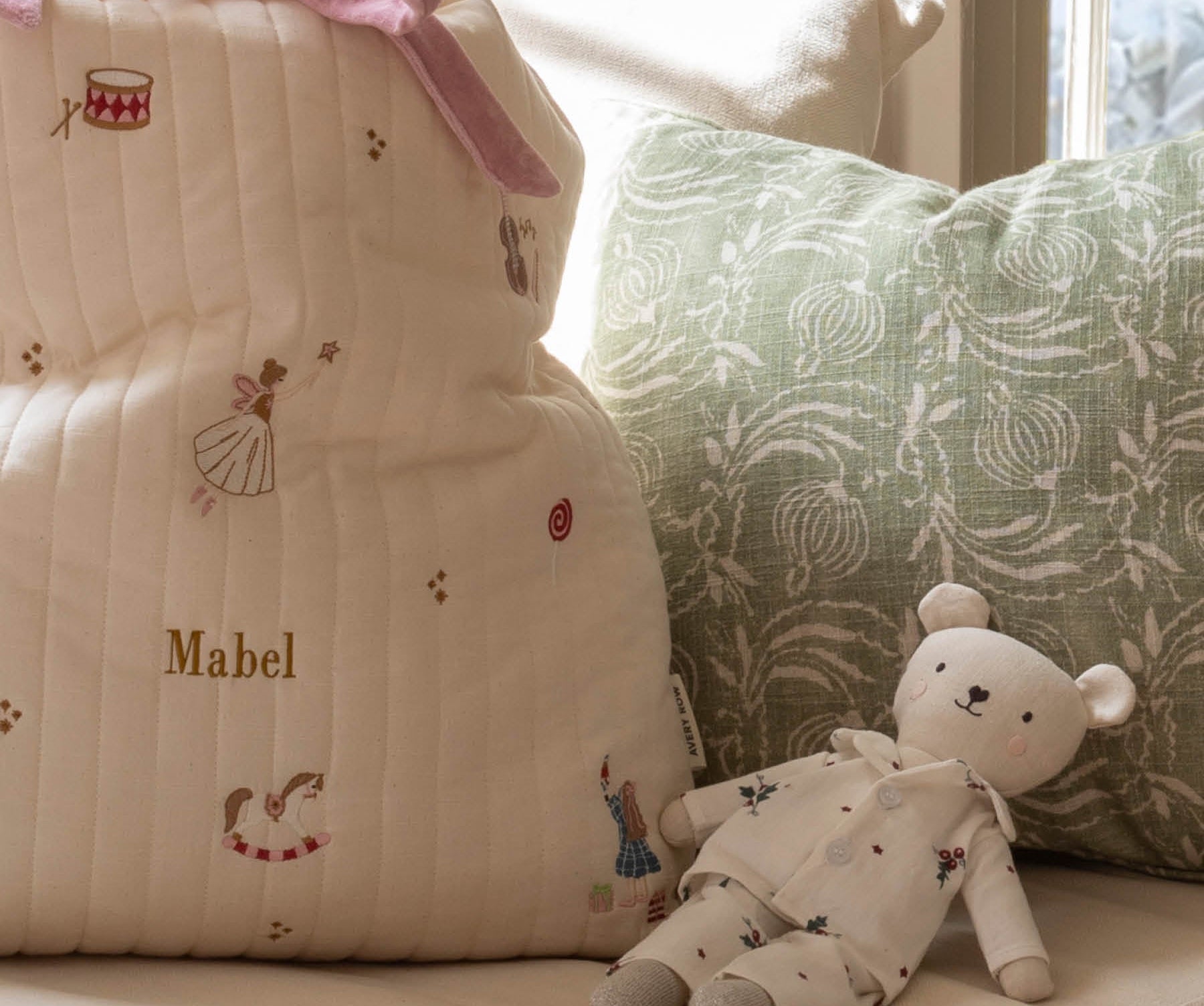 A cream quilted baby bag with embroidered ballerina and bird motifs, personalized with the name 'Mabel' in gold, styled on a sofa next to a green patterned pillow and a teddy bear.