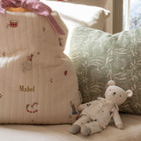 A cream quilted baby bag with embroidered ballerina and bird motifs, personalized with the name 'Mabel' in gold, styled on a sofa next to a green patterned pillow and a teddy bear.