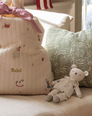 A cream quilted baby bag with embroidered ballerina and bird motifs, personalized with the name 'Mabel' in gold, styled on a sofa next to a green patterned pillow and a teddy bear.