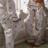 Three children wearing matching white long-sleeve pajamas with a small red and green floral print and red piping trim, standing barefoot indoors near a window.