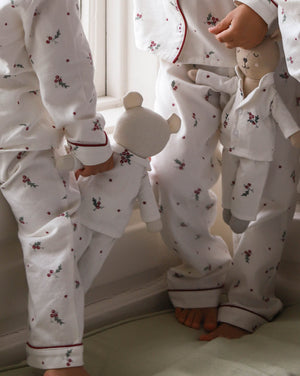 Three children wearing matching white long-sleeve pajamas with a small red and green floral print and red piping trim, standing barefoot indoors near a window.