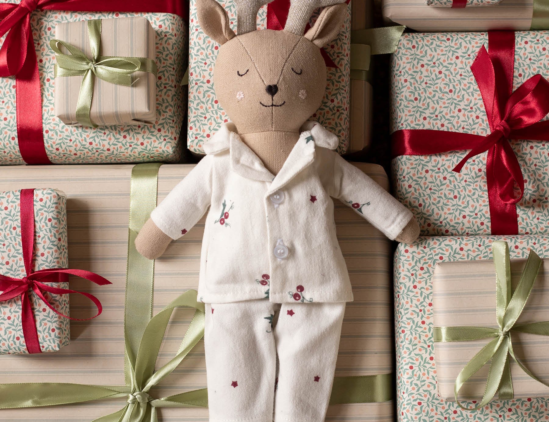 A plush deer doll wearing cream pajamas with small red floral embroidery, posed among wrapped gift boxes with red and green ribbons and patterned paper.