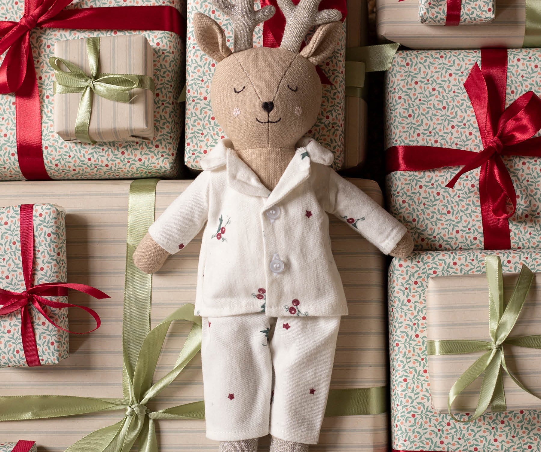 A plush deer doll wearing cream pajamas with small red floral embroidery, posed among wrapped gift boxes with red and green ribbons and patterned paper.