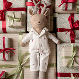A plush deer doll wearing cream pajamas with small red floral embroidery, posed among wrapped gift boxes with red and green ribbons and patterned paper.
