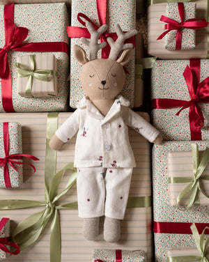 A plush deer doll wearing cream pajamas with small red floral embroidery, posed among wrapped gift boxes with red and green ribbons and patterned paper.