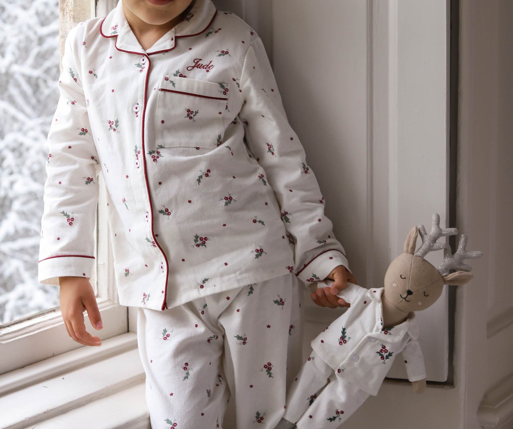 A young child wearing a white long-sleeve pajama set with red piping and a small floral print, standing by a window and holding a small doll dressed in matching pajamas.