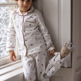 A young child wearing a white long-sleeve pajama set with red piping and a small floral print, standing by a window and holding a small doll dressed in matching pajamas.