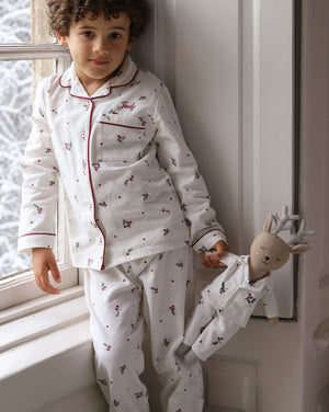 A young child wearing a white long-sleeve pajama set with red piping and a small floral print, standing by a window and holding a small doll dressed in matching pajamas.