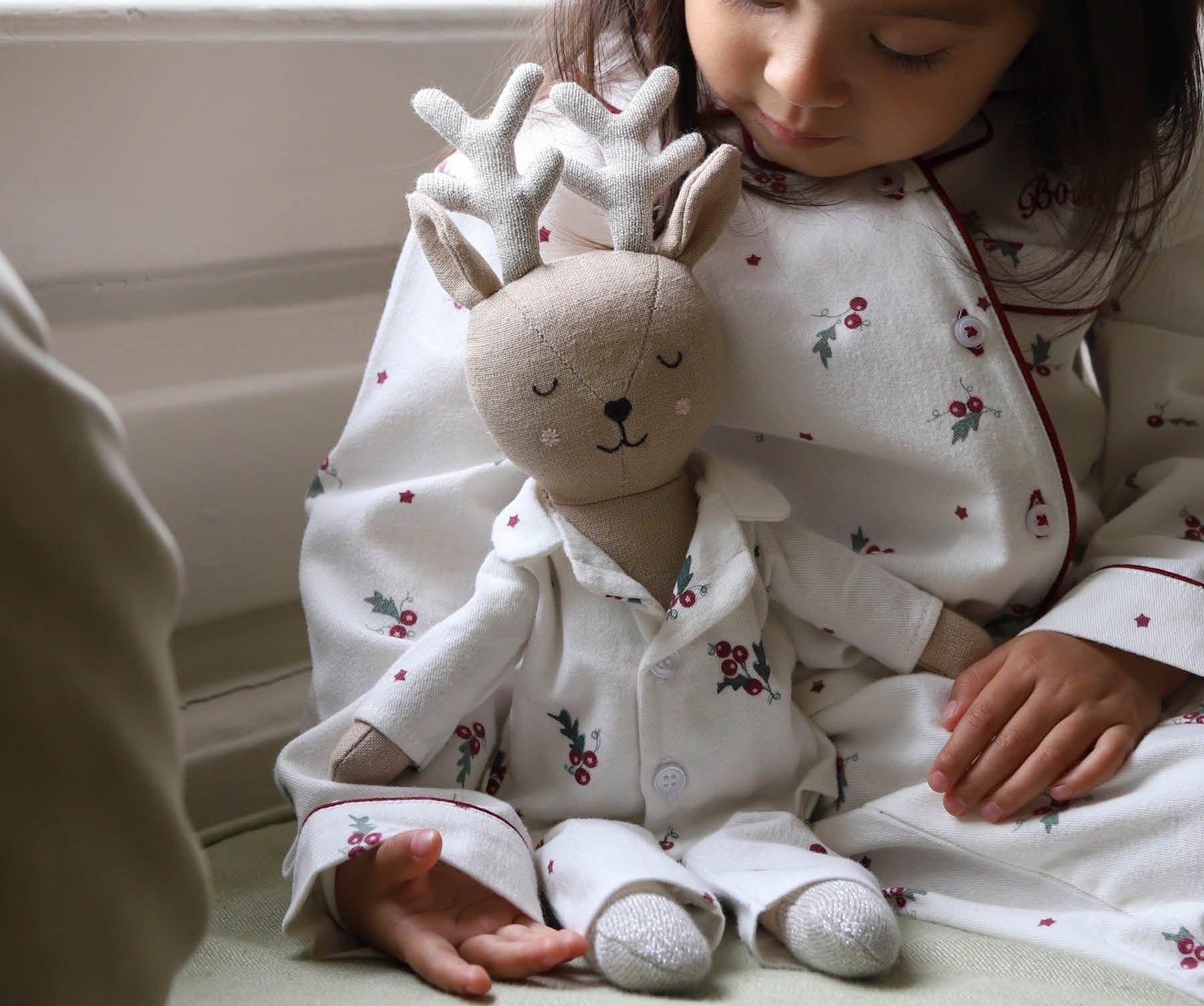 A young child wearing white Christmas-themed pajamas sits indoors next to a matching plush stuffed toy dressed as a reindeer, with soft natural light coming from a nearby window.