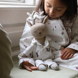 A young child wearing white Christmas-themed pajamas sits indoors next to a matching plush stuffed toy dressed as a reindeer, with soft natural light coming from a nearby window.
