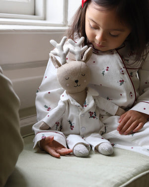 A young child wearing white Christmas-themed pajamas sits indoors next to a matching plush stuffed toy dressed as a reindeer, with soft natural light coming from a nearby window.