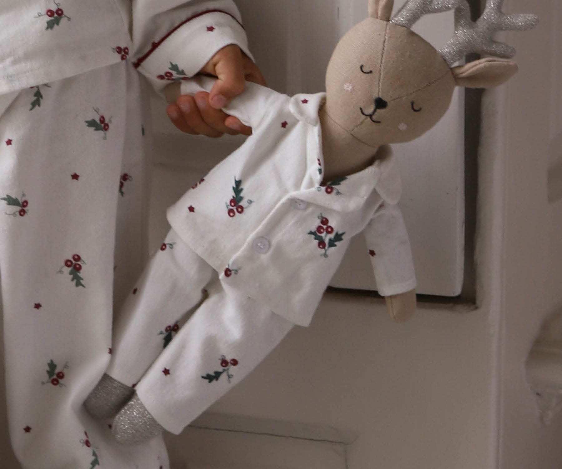 A child wearing white holiday-themed pajamas with a red berry and holly print, holding a plush stuffed deer doll dressed in matching white pajamas with glittery silver antlers.