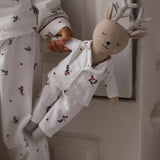 A child wearing white holiday-themed pajamas with a red berry and holly print, holding a plush stuffed deer doll dressed in matching white pajamas with glittery silver antlers.