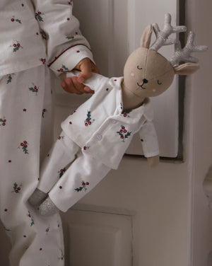 A child wearing white holiday-themed pajamas with a red berry and holly print, holding a plush stuffed deer doll dressed in matching white pajamas with glittery silver antlers.