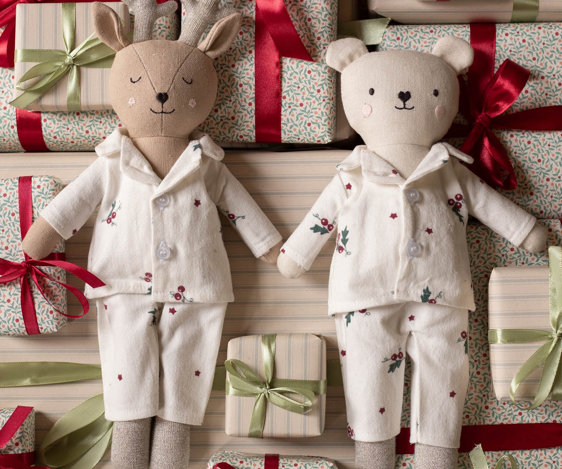 Two plush stuffed toys dressed in matching holiday pajamas, surrounded by wrapped gift boxes with red and green ribbons. The left toy is a reindeer with antlers, and the right toy is a bear, both in cream pajamas with small floral and star prints.