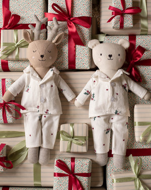 Two plush stuffed toys dressed in matching holiday pajamas, surrounded by wrapped gift boxes with red and green ribbons. The left toy is a reindeer with antlers, and the right toy is a bear, both in cream pajamas with small floral and star prints.