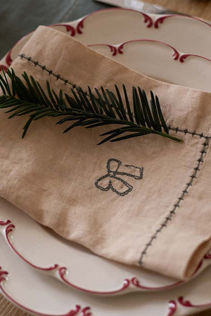 Folded beige napkin with embroidery on a set of white plates with red trim.