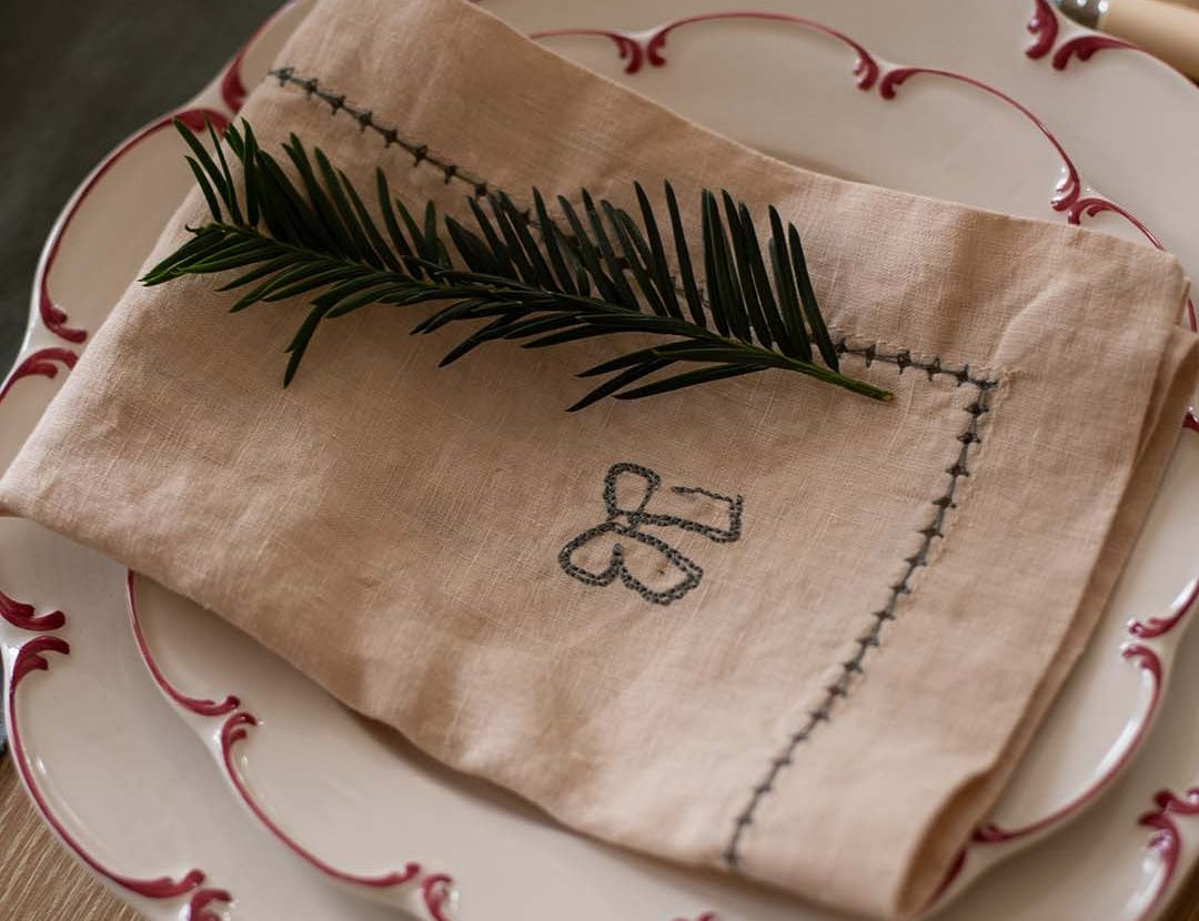 Folded beige napkin with embroidery on a set of white plates with red trim.
