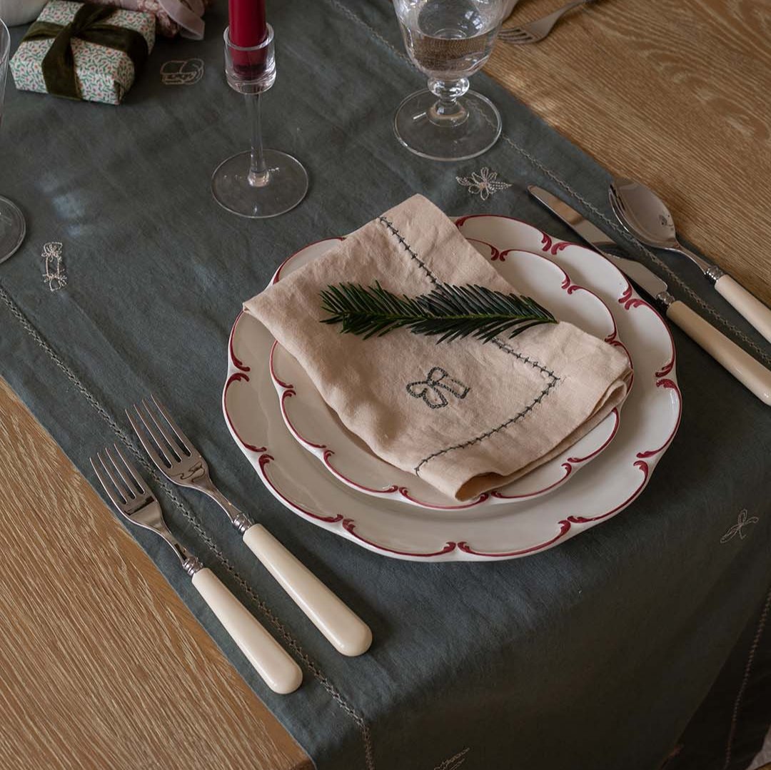 A styled table setting featuring a dark gray tablecloth with embroidered details, layered white plates with red scalloped edges, a beige embroidered napkin with a green pine branch on top, and cream-handled cutlery on a wooden table.