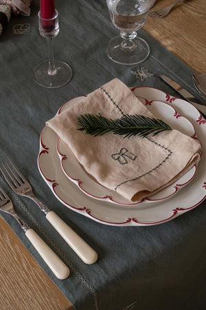 A styled table setting featuring a dark gray tablecloth with embroidered details, layered white plates with red scalloped edges, a beige embroidered napkin with a green pine branch on top, and cream-handled cutlery on a wooden table.