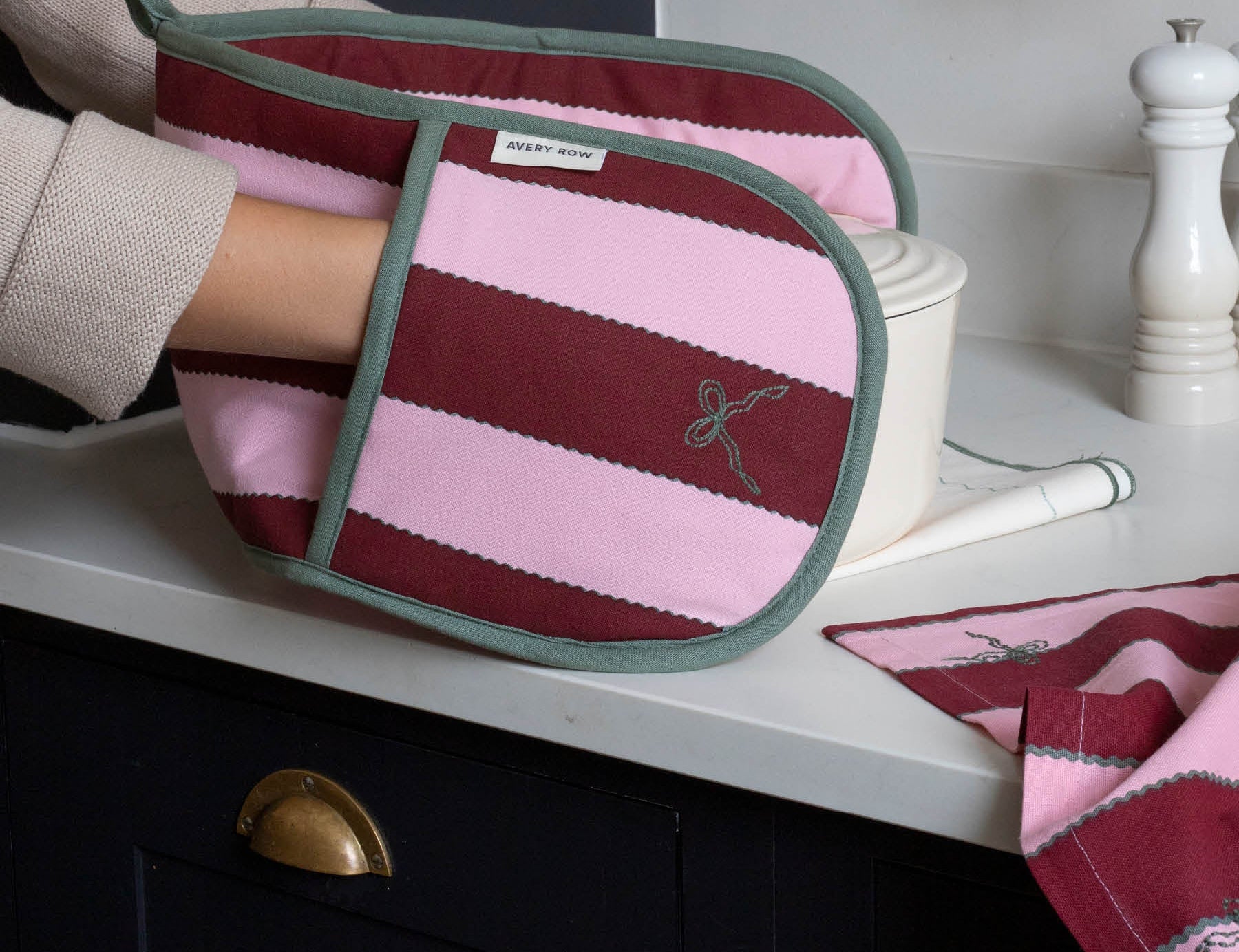 A striped fabric oven mitt with pink and burgundy horizontal stripes, sage green trim, and a small embroidered bow motif, being held by a person in a kitchen setting.