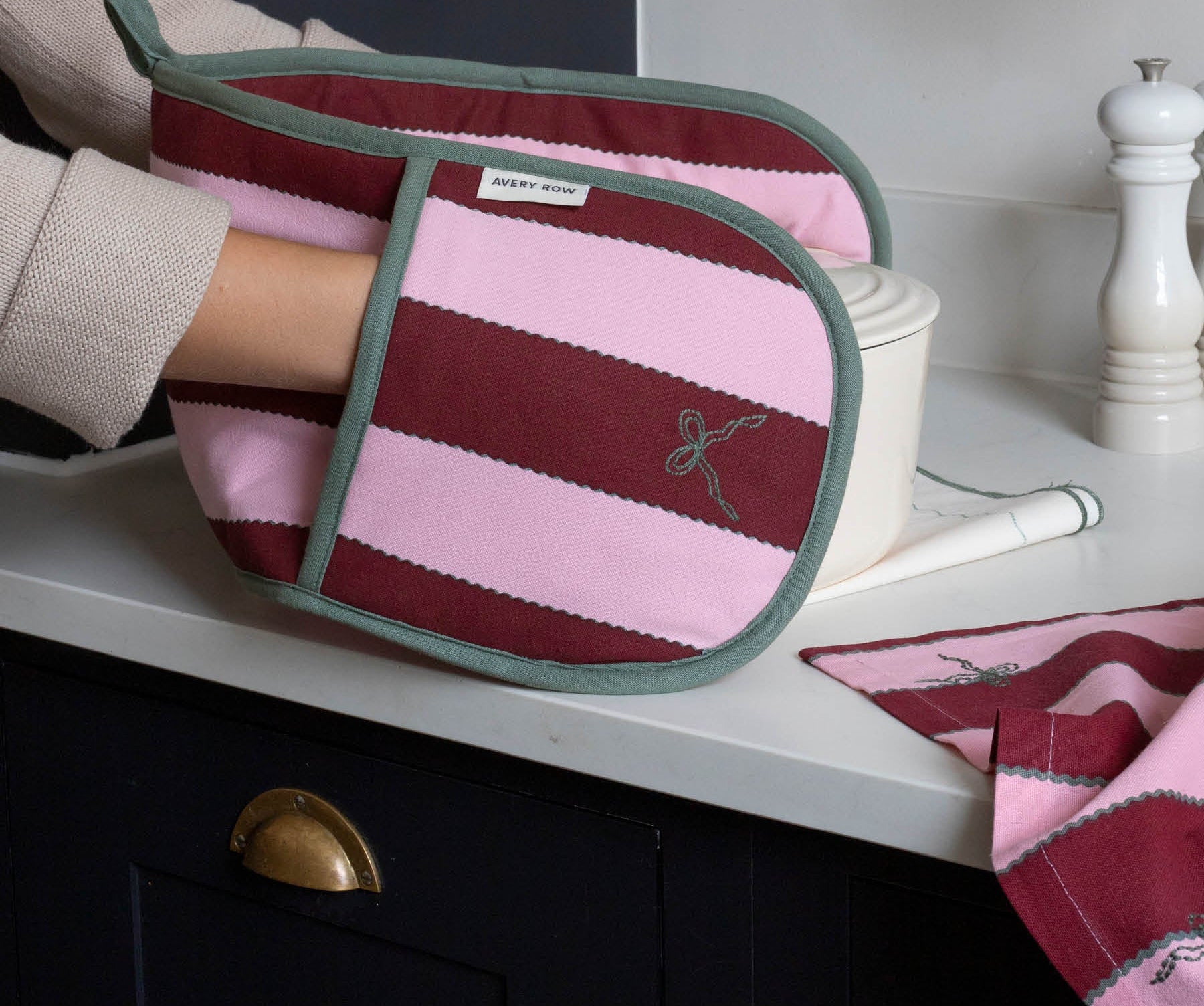 A striped fabric oven mitt with pink and burgundy horizontal stripes, sage green trim, and a small embroidered bow motif, being held by a person in a kitchen setting.