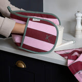A striped fabric oven mitt with pink and burgundy horizontal stripes, sage green trim, and a small embroidered bow motif, being held by a person in a kitchen setting.