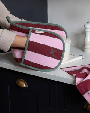 A striped fabric oven mitt with pink and burgundy horizontal stripes, sage green trim, and a small embroidered bow motif, being held by a person in a kitchen setting.