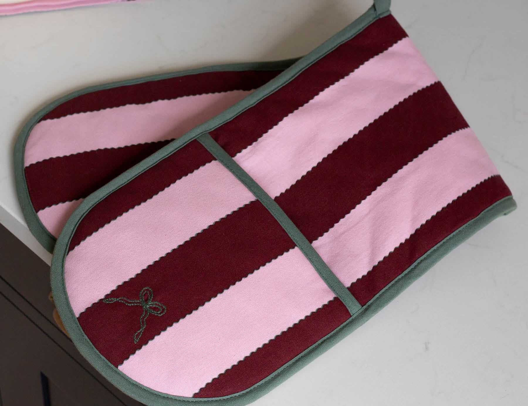 A pair of striped oven mitts with horizontal bands of dusty pink and deep burgundy, finished with sage green trim and handles, resting on a white countertop.