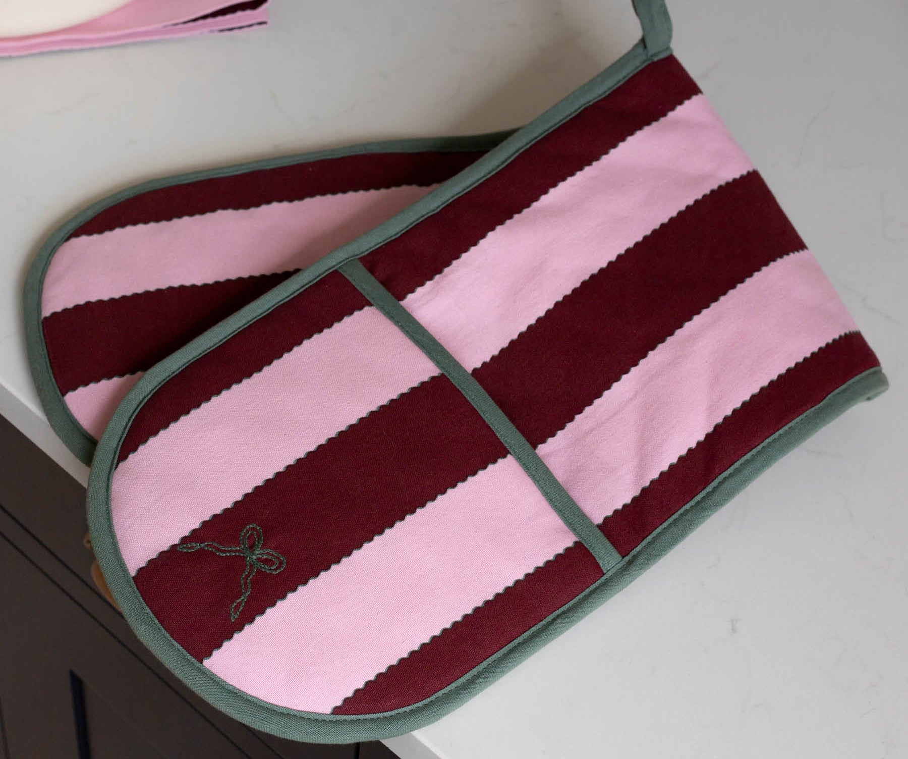 A pair of striped oven mitts with horizontal bands of dusty pink and deep burgundy, finished with sage green trim and handles, resting on a white countertop.