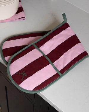 A pair of striped oven mitts with horizontal bands of dusty pink and deep burgundy, finished with sage green trim and handles, resting on a white countertop.