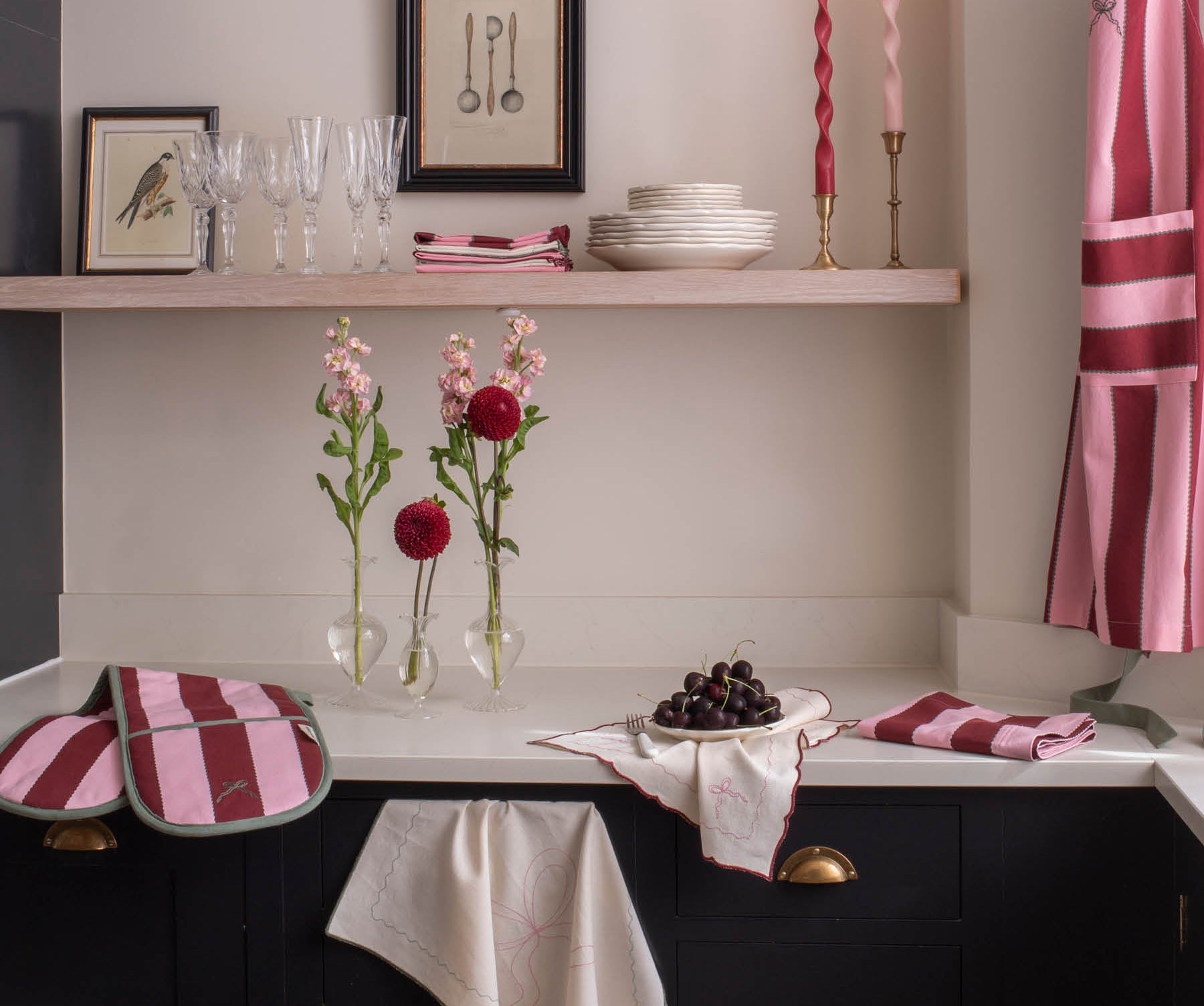 A styled kitchen corner with dark cabinetry, a white countertop, pink striped textiles, framed wall art, glassware, candles, and flowers. No person or animal is present, and the scene appears to be a real interior environment rather than a clean isolated product shot, so this fits Lifestyle rather than Product/Packaging.