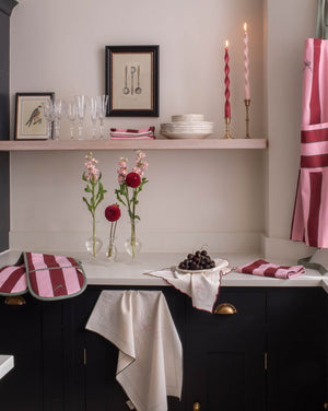 A styled kitchen corner with dark cabinetry, a white countertop, pink striped textiles, framed wall art, glassware, candles, and flowers. No person or animal is present, and the scene appears to be a real interior environment rather than a clean isolated product shot, so this fits Lifestyle rather than Product/Packaging.